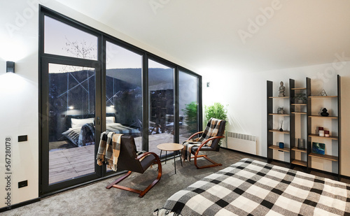 Bedroom with comfortable furniture and irregular panoramic window at twilight. Cozy modern furnished cottage room with reflecting in large curtainless window in evening. Concept of interior. photo