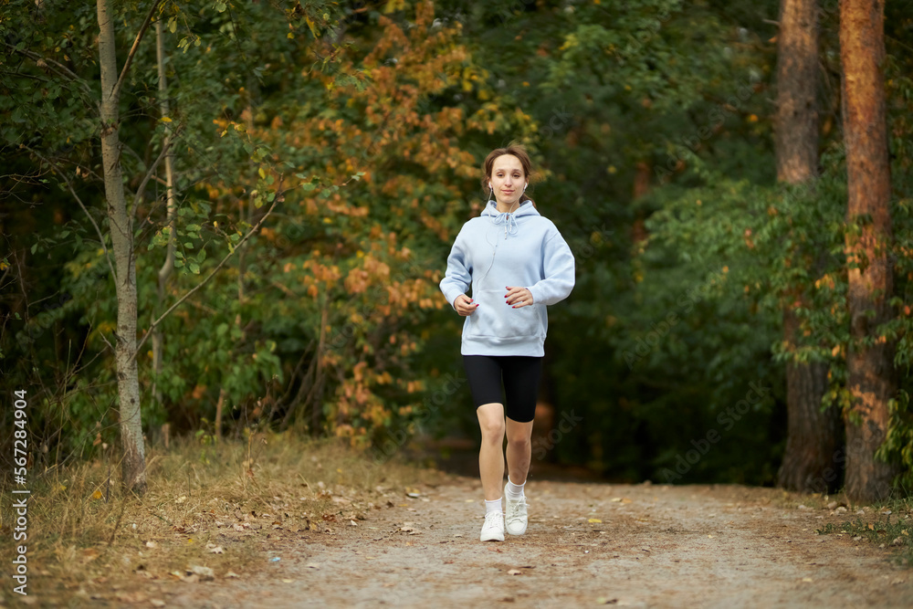 a caucasian girl in a blue sweatshirt puts on headphones in the park and prepares to run. pretty young woman puts on headphones. Pretty smiling woman puts on headphones in the park before jogging. a caucasian girl in a blue sweatshirt puts on headphones in the park and prepares to run. pretty young woman puts on headphones. Pretty smiling woman puts on headphones in the park before jogging.