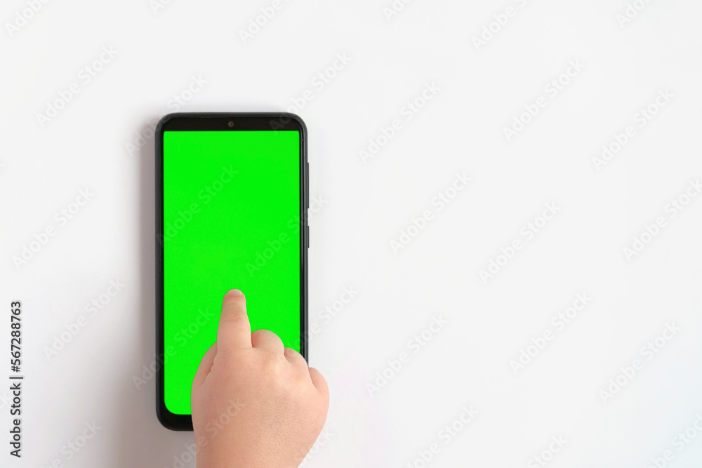 Child hand touching clicking on mobile phone with chromakey screen. Little Baby Finger Taps in the center of blank phone green display. Close up. Using smartphone device. Top view Flat lay Child hand touching clicking on mobile phone with chromakey screen. Little Baby Finger Taps in the center of blank phone green display. Close up. Using smartphone device. Top view Flat lay