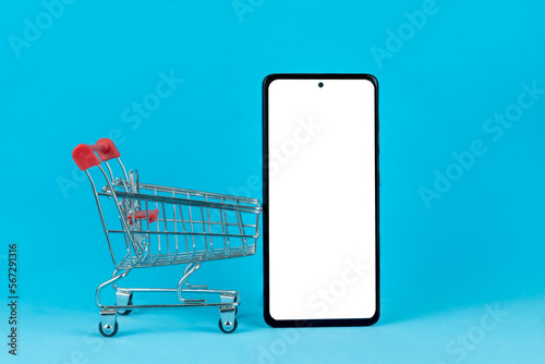 Shopping cart with a nearby mobile phone with a white screen on a blue background, copy the space. Online shopping concept, delivery