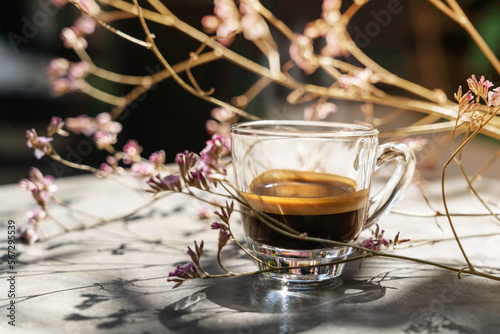 Espresso shot coffee in clear small cup with smoke above and blur pink flower in background in the morning
