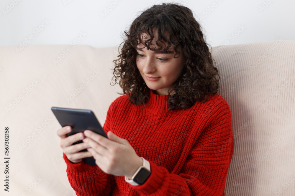 Curly white girl browsing internet on tablet computer at home. Portrait of dark haired young female reading ebook online Curly white girl browsing internet on tablet computer at home. Portrait of dark haired young female reading ebook online