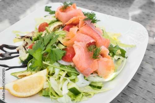 A smoked salmon and avocado salad with green lettuce, dill, cherry tomatoes and cucumber, with a slice of lemon