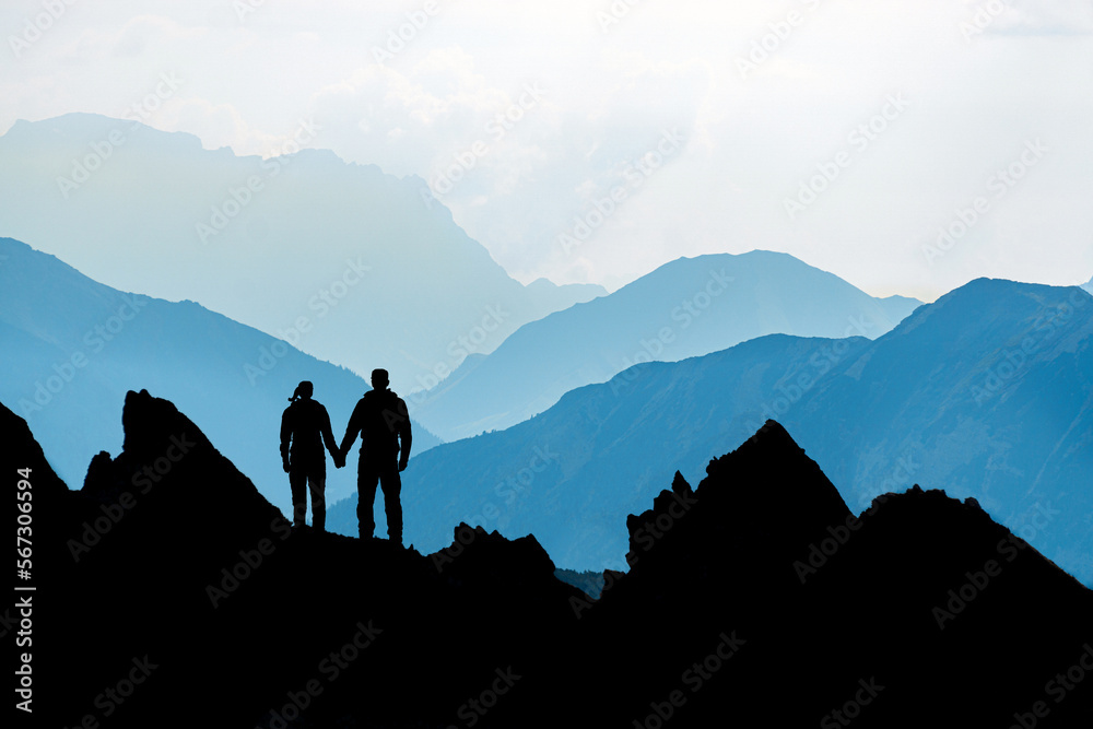Silhouette Couple of man and woman reaching mountain top enjoying freedom and looking towards blue mountain silhouettes and sunrise. Alps, Allgaeu, Bavaria, Germany.