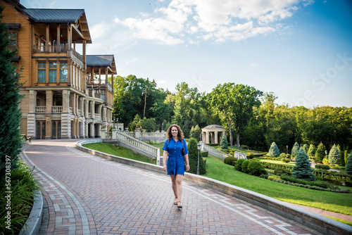 Beautiful young smiling woman walking in city park near famous old architecture building at summer day wearing dress and looking to the camera