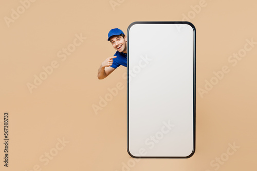 Full body delivery guy employee man wear blue cap t-shirt uniform workwear work as dealer courier stand behind point on big huge blank screen area mobile cell phone isolated on plain beige background. photo