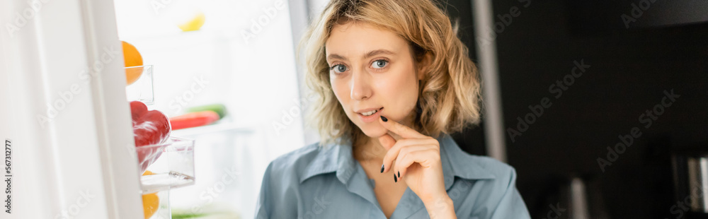 pensive young woman looking at camera near open refrigerator in kitchen, banner. pensive young woman looking at camera near open refrigerator in kitchen, banner.