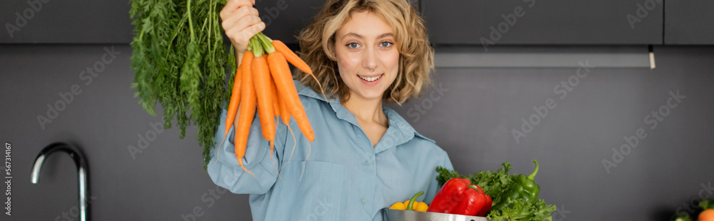 cheerful young woman holding bowl with vegetables and fresh carrots in kitchen, banner. cheerful young woman holding bowl with vegetables and fresh carrots in kitchen, banner.