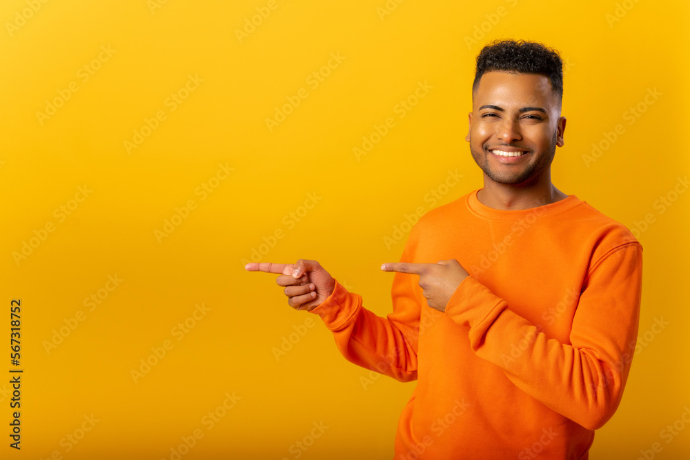 Portrait of pleasant looking indian man wearing orange pullover pointing fingers aside at copy space, showing area for advertisement. Indoor studio shot isolated on yellow background Portrait of pleasant looking indian man wearing orange pullover pointing fingers aside at copy space, showing area for advertisement. Indoor studio shot isolated on yellow background