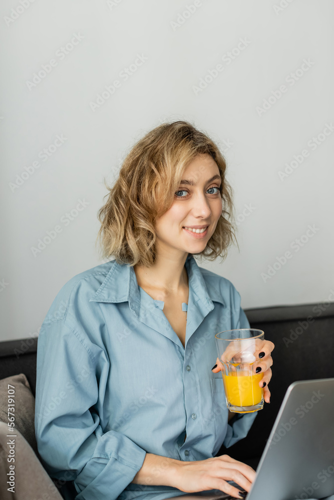 smiling young freelancer with wavy hair holding glass of orange juice near laptop. smiling young freelancer with wavy hair holding glass of orange juice near laptop.
