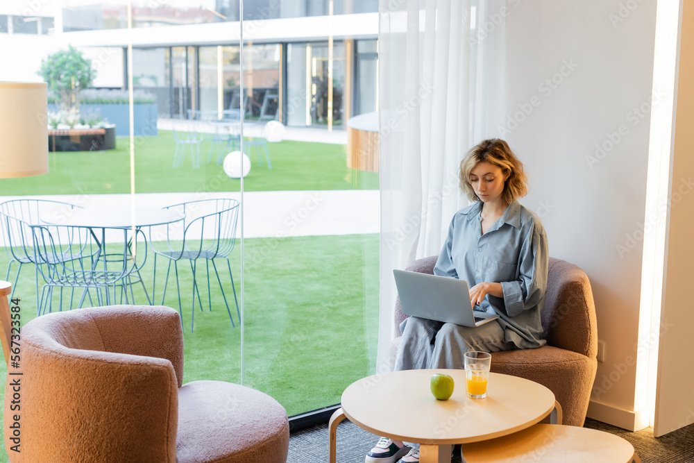 young blonde woman with wavy hair using laptop while sitting on armchair in lobby of hotel. young blonde woman with wavy hair using laptop while sitting on armchair in lobby of hotel.