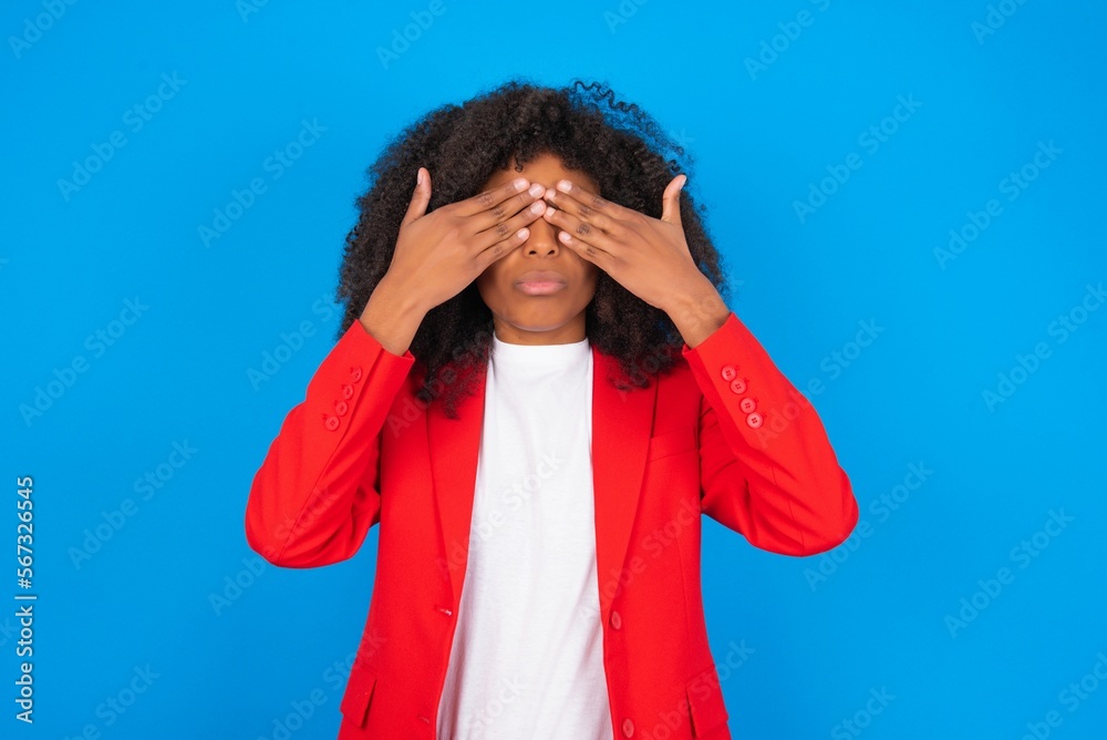 young businesswoman with afro hairstyle wearing red over blue background covering eyes with both hands, doesn't want to see anything or feeling ashamed. Human feelings reactions. young businesswoman with afro hairstyle wearing red over blue background covering eyes with both hands, doesn't want to see anything or feeling ashamed. Human feelings reactions.
