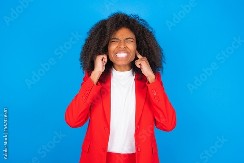 Tablou pe pânză Stop making this annoying sound! Unhappy stressed out young businesswoman with afro hairstyle wearing red over blue background making worry face, plugging ears with fingers, irritated with loud noise