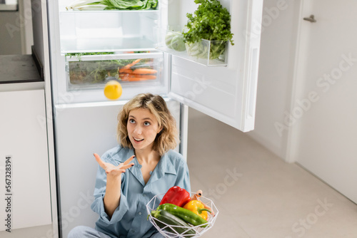 young woman sitting near opened refrigerator with fresh vegetables while throwing lemon in air.