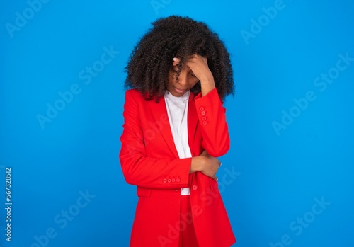 young businesswoman with afro hairstyle wearing red over blue background making facepalm gesture while smiling amazed with stupid situation. photo