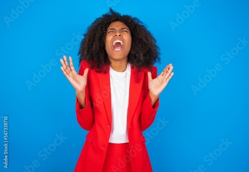 Crazy outraged young businesswoman with afro hairstyle wearing red over blue background screams loudly and gestures angrily yells furiously. Negative human emotions feelings concept photo