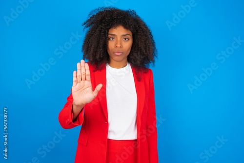 young businesswoman with afro hairstyle wearing red over blue background shows stop sign prohibition symbol keeps palm forward to camera with strict expression photo