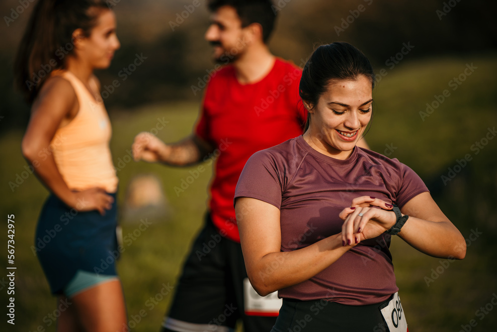 Caucasian woman runner set the smartwatch before running a mountain trail at sunset. Two people in the background.