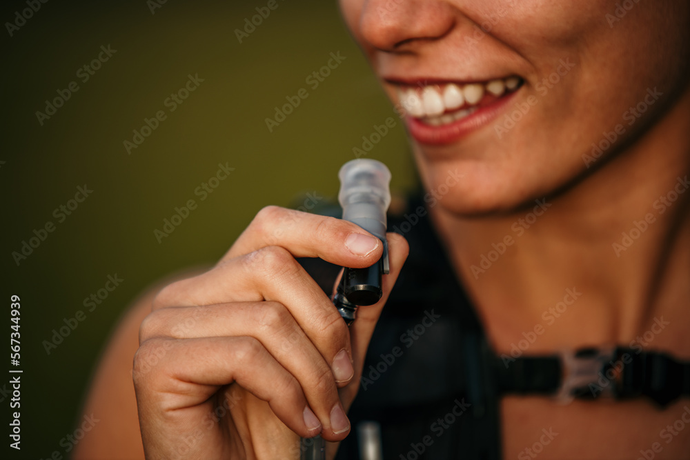 A smiling female trail runner is about to drink water from hydration packs and rest.