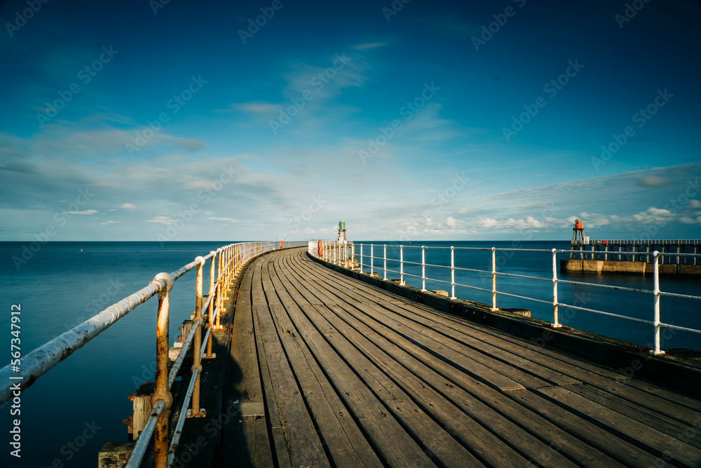 Obraz premium Whitby Harbour pier with lighthouse and wooden boards with metal railings and the sea in the background.