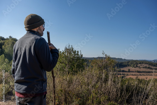 man in his forties with a walking stick, hat and clothes for the cold, looking at the landscape, looking like a shepherd and a country man. Beard and glasses. Mediterranean forest. blue sky.