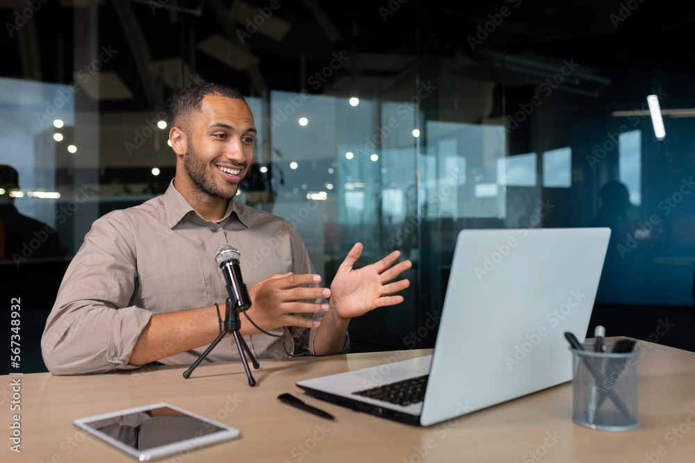 Successful hispanic man inside office with professional microphone recording audiobook and online podcast, business coach work with laptop live event Businessman in casual shirt glasses at workplace