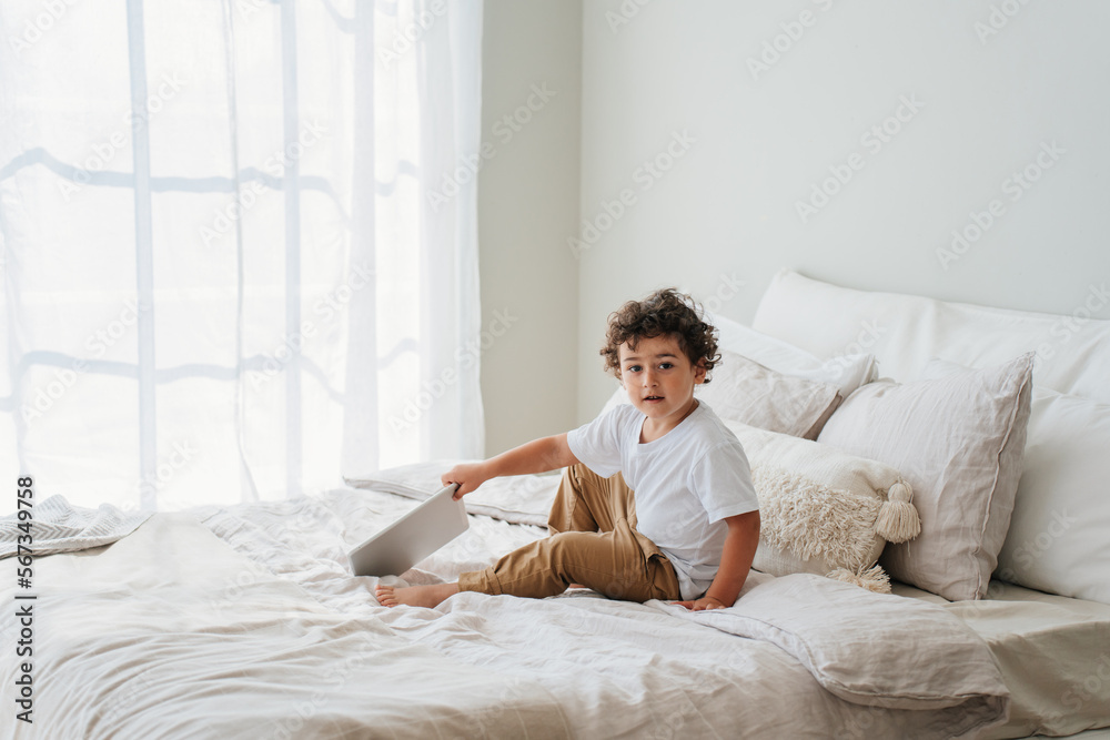 Handsome curly caucasian baby boy sitting on bed holds tablet looks at camera on sunny day. Childhood and gadgets. Smart Hispanic little kid enjoys games via internet. Cute toddler plays home. Handsome curly caucasian baby boy sitting on bed holds tablet looks at camera on sunny day. Childhood and gadgets. Smart Hispanic little kid enjoys games via internet. Cute toddler plays home.