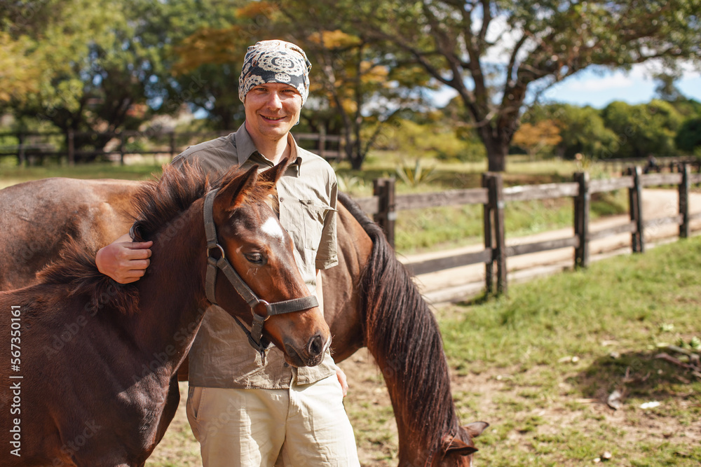 Smiling, young man standing next to brown horse foal, another animal in background Smiling, young man standing next to brown horse foal, another animal in background