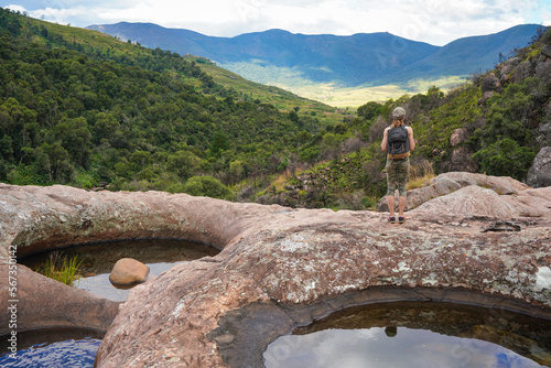 Young woman in hiking clothes, standing on stones near natural water pool, blurred trees and mountains background, view form behind - typical scenery seen during trek in Andringitra, Madagascar photo