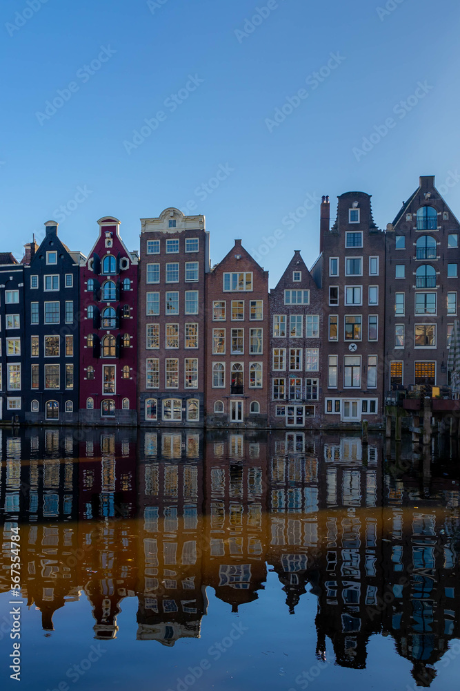 Architecture features traditional houses with blue sky and reflection on water, Typical dutch houses at Damrak canal, An avenue between Amsterdam Centraal and Dam Square, Noord Holland, Netherlands.