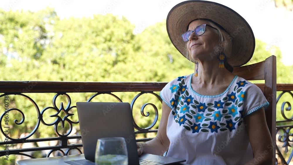Fototapeta premium Portrait of pretty smiling happy mature woman digital nomad wearing ethnic blouse and straw hat using a laptop computer on a balcony in Europe with trees blurry in background.
