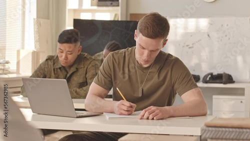 Waist up slowmo of group of young multiethnic male and female cadets sitting at their desks in classroom and studying military service with professor at academy