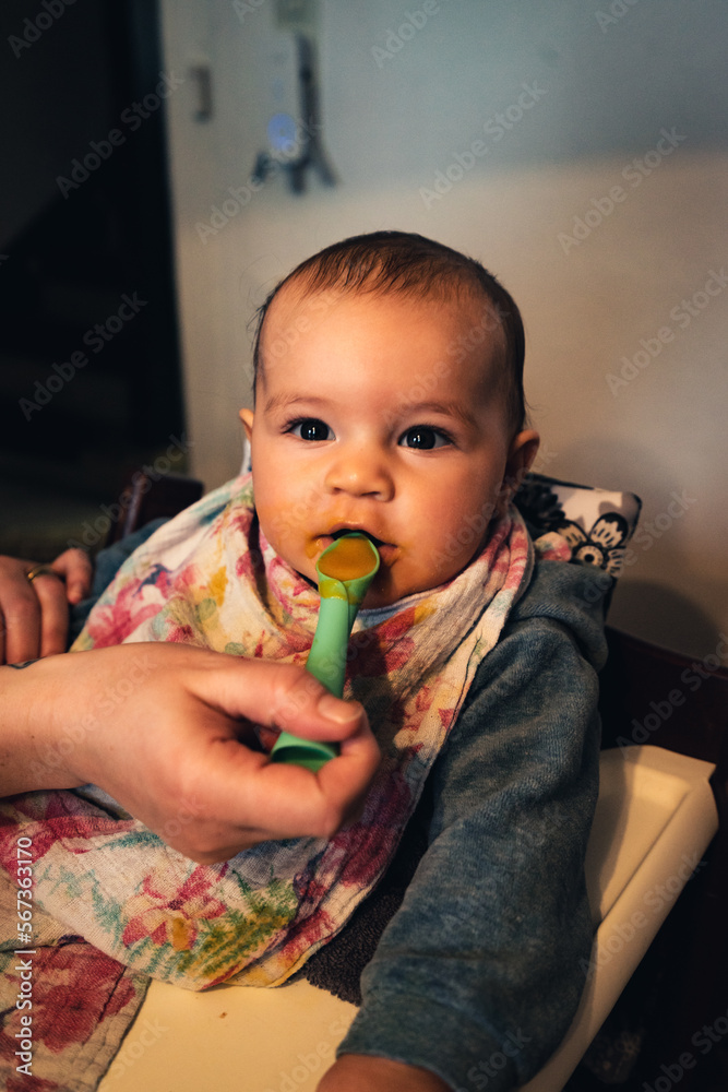 Baby mit großem bunten Lätzchen und verschmiertem Gesicht in einem Kindersitz wird gefüttert Baby mit großem bunten Lätzchen und verschmiertem Gesicht in einem Kindersitz wird gefüttert