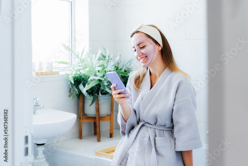 Smiling woman with facial pink clay mask on the face using smartphone at home bath enjoying relaxation and spa beauty treatment. Teen Beauty blogger creating content. Selective focus, copy space