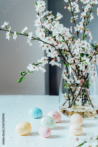 Photography Blooming tree branches in the vase and colored easter eggs on the white kitchen table with simple interior background