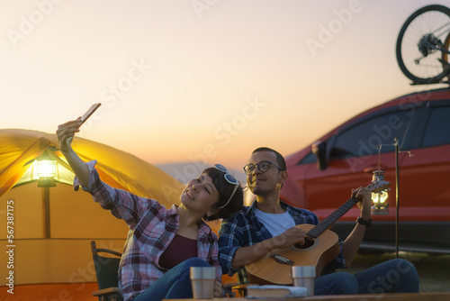 Asian couple singing and playing guitar while taking a selfie by smartphone camera in their camping area with lake in the background during sunset.