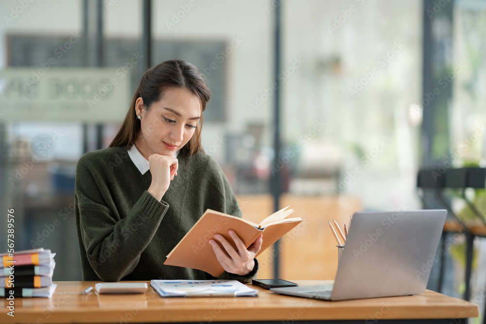 Beautiful Asian businesswoman working on a laptop and having fun at work and reading review book documenting detailed financial information earning peace of mind.