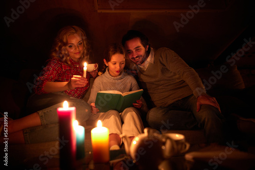 Cozy family evening. Mother, father and little girl sitting on sofa without electricity and reading book with candle light. Blackout. Hobby, leisure time. Concept of power outage, adjusting