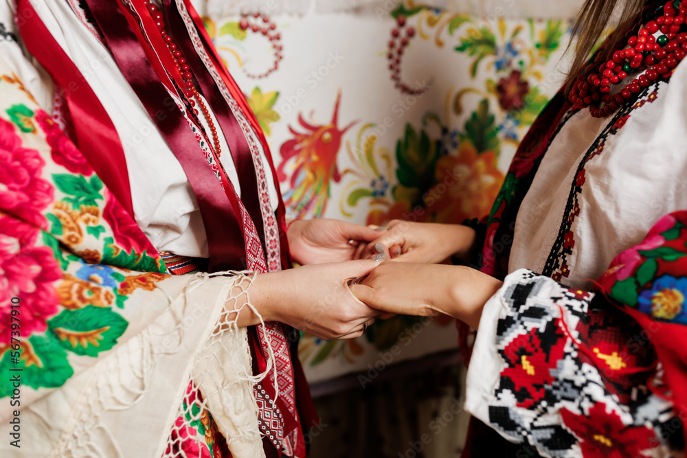 Two ukrainian women in traditional ethnic clothing holding hands on background of decorated stove in hut