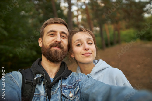 A caucasian couple in love takes a selfie in the park, against the backdrop of trees. A guy and a girl take a selfie close-up. A guy in a denim jacket and a girl in a hoodie take a selfie