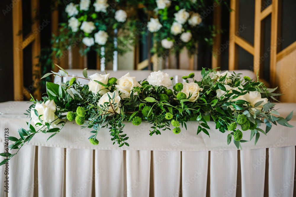 Festive table with tablecloth near arch, decorated with composition of flowers and greenery, candles in the banquet hall. Table newlyweds in the banquet area on party in restaurant. Wedding setup.