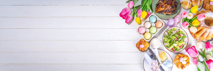 Festive served Easter dinner or brunch table, with Easter egg, tulip flower bouquet, traditional Easter foods - cake, cross buns, wreath bread, glazed ham and spring salad, white table background