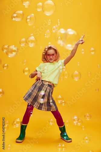 Beautiful girl, child in stylish clothes, skirt and rubber boots, dancing, posing over yellow studio background with soap bubbles. Concept of childhood, emotions, fun, fashion, facial expression