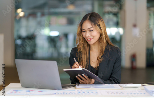 Beautiful Asian businesswoman using her laptop to work and enjoy taking notes on meeting minutes agendas of secretarial work.
