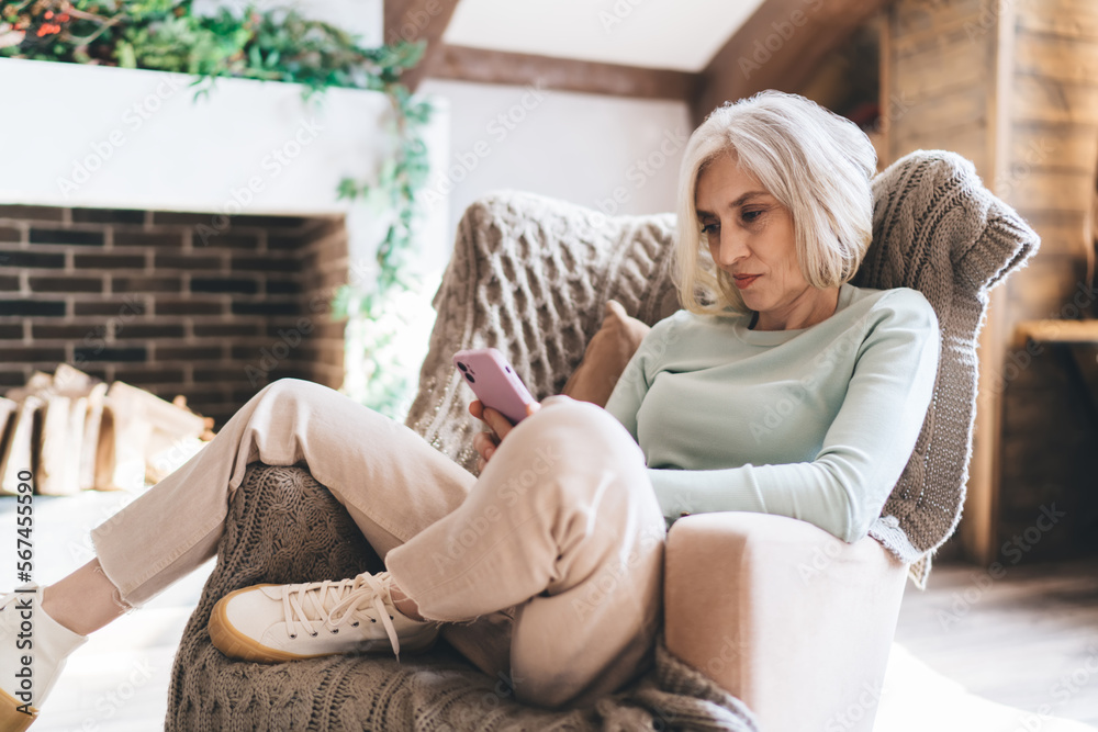 Obraz premium Mature woman browsing smartphone while sitting on sofa