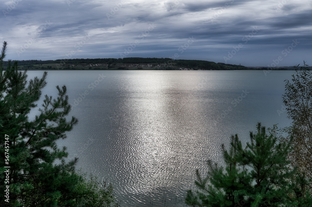 Pine trees grow on the banks of a large river, a beautiful cloudy sky, the surface of the water reflects sunlight with light ripples, a beautiful summer landscape