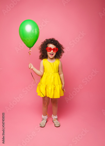 Happy birthday celebration with flying balloons of a charming cute little girl in a yellow dress isolated on a pink background. © inna717