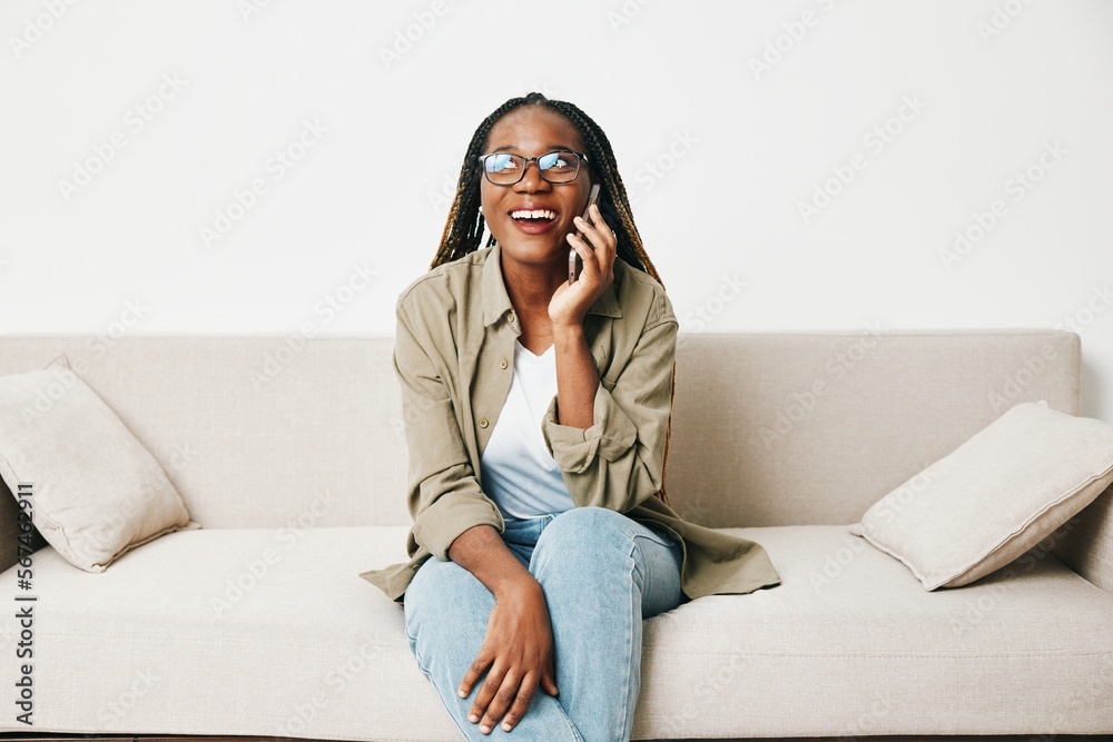 African American woman business freelancer working sitting on the couch at home in the phone, business calls and messages happiness smile, home clothes and eyeglasses, light interior background.