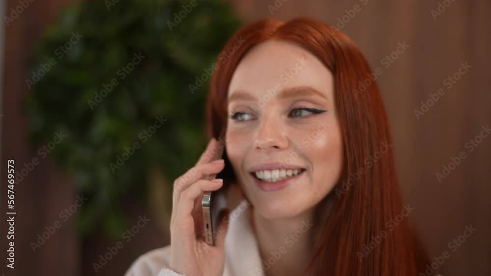 Close-up face of cheerful young woman wearing white bathrobe talking using mobile smartphone while resting at home. Close up portrait of smiling redhead female having phone call after spa treatments. Close-up face of cheerful young woman wearing white bathrobe talking using mobile smartphone while resting at home. Close up portrait of smiling redhead female having phone call after spa treatments.