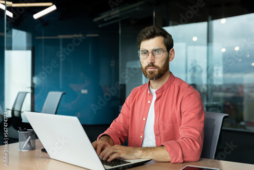 Serious and focused mature businessman in red shirt wearing glasses looking at camera, young man at work typing on laptop keyboard, programmer developer portrait at work.